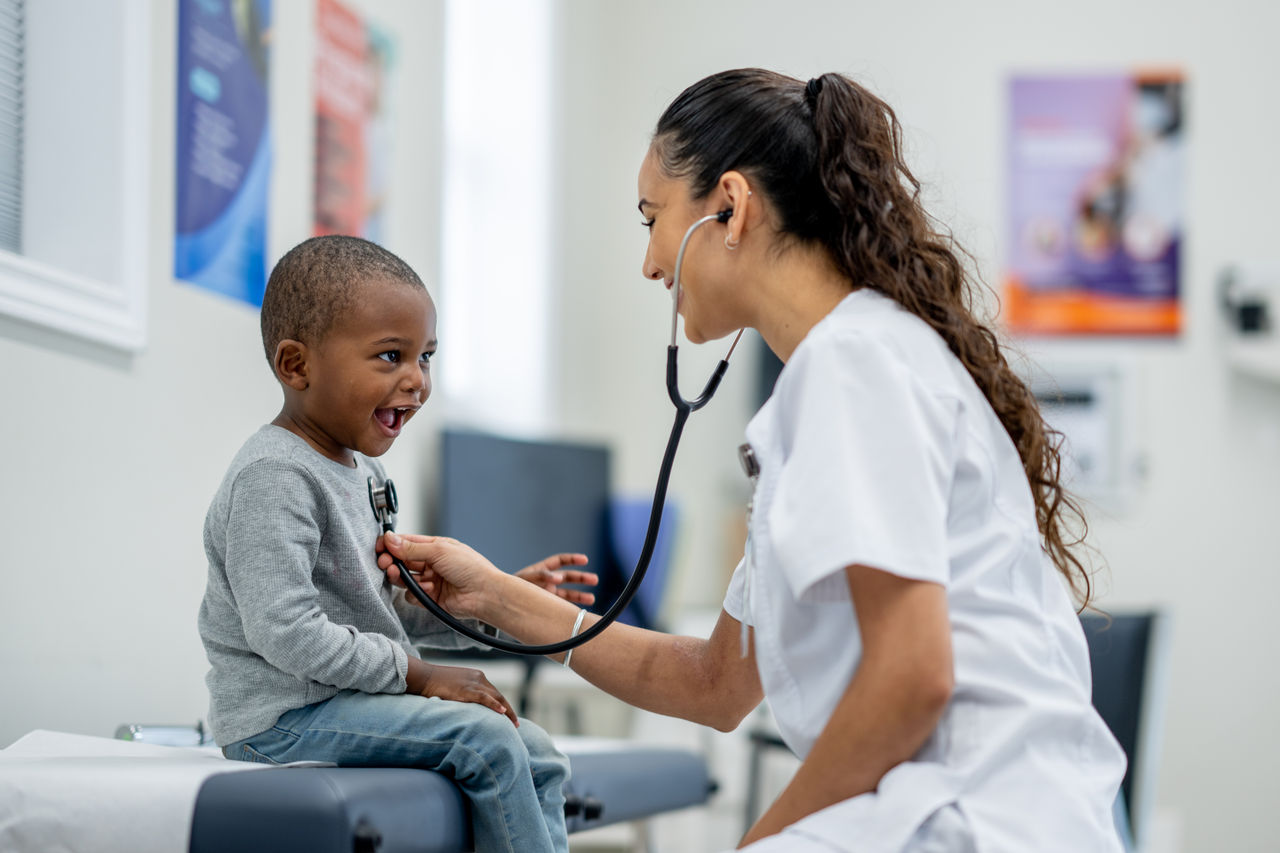 A sweet little boy sits up on an exam table in a doctors office during a routine check-up.  His female doctor is seated in front of him and listening to his heart with her stethoscope.